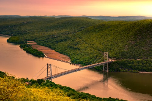 A scenic view of a bridge spanning a river, surrounded by lush green hills at sunset.