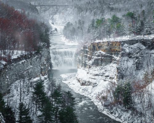 A snowy landscape featuring a waterfall cascading into a river, surrounded by cliffs and evergreen trees.