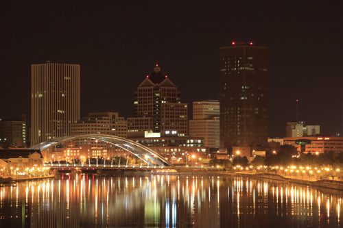 City skyline at night with illuminated buildings and a bridge reflecting in the water.
