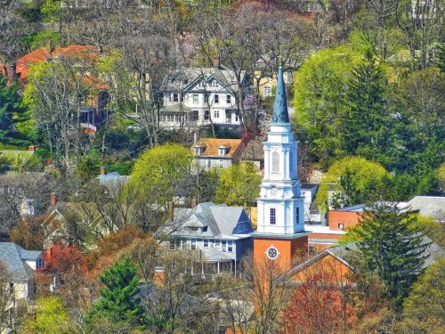 A scenic view of a church steeple surrounded by trees and historic homes in a hilly landscape during spring.