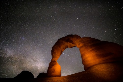 A night sky filled with stars above a glowing rock arch, with a faint meteor streaking across the scene.