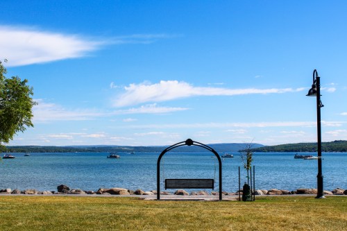 A scenic view of a lake with boats, framed by a bench and a lamp post under a clear blue sky.