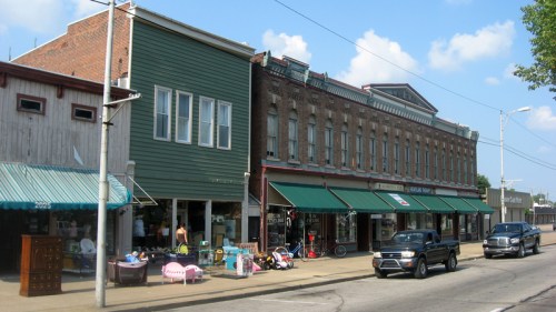 Historic storefronts line a street, featuring a mix of brick and wooden buildings with awnings and parked cars.