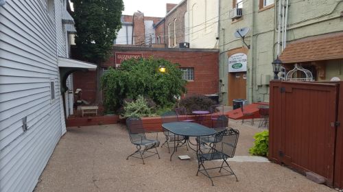 A small outdoor patio with tables and chairs, surrounded by greenery and brick walls, in a cozy urban setting.