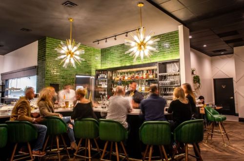 A lively bar scene with patrons at a green-tiled counter, stylish lighting, and a well-stocked bar in the background.