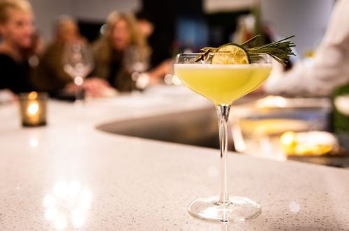 A cocktail in a glass garnished with lime and rosemary, placed on a bar with blurred patrons in the background.
