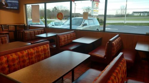 Interior of a fast-food restaurant featuring empty booths and tables, with large windows showing a parking lot outside.