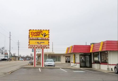 Sign for Penguin Point Drive-Thru restaurant with a red roof and a parking lot, set against a cloudy sky.