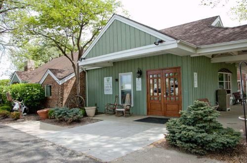 A green building with a wooden entrance, surrounded by trees and shrubs, featuring outdoor seating and a welcoming atmosphere.
