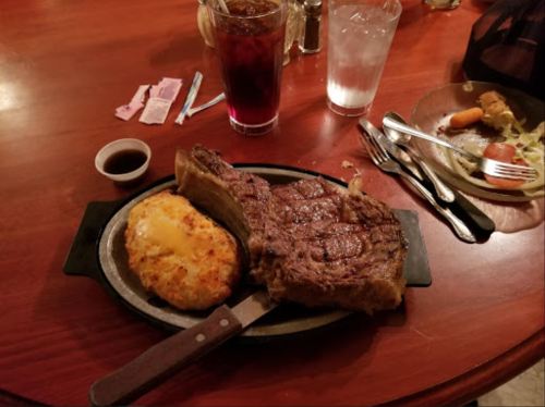 A large ribeye steak on a sizzling plate with cheesy potatoes, accompanied by drinks and a partially eaten side salad.