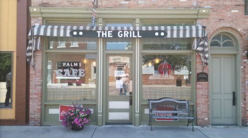 Front view of a cafe named "The Grill," featuring a striped awning, flower pots, and a bench outside.