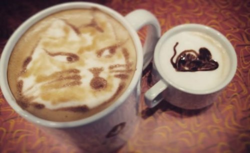 A large coffee cup with a cat face design in the foam, next to a small cup with a heart shape in the foam.