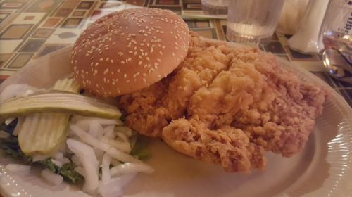 A plate featuring a sesame seed bun, crispy fried chicken, pickles, and sliced onions on a bed of lettuce.