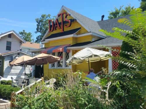 A bright yellow restaurant with a sign reading "DATS," featuring outdoor seating and greenery in the foreground.