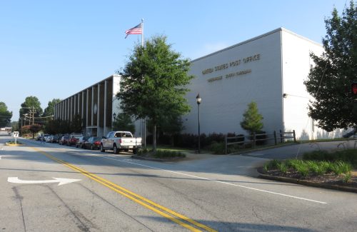 A U.S. Post Office building in a small town, with an American flag and parked cars along the street.