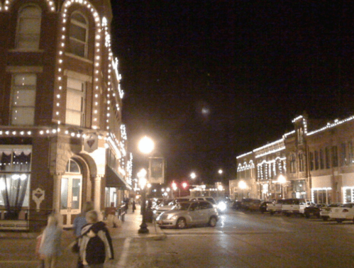 A charming street at night, lined with historic buildings adorned with string lights and parked cars.