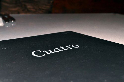 A close-up of a black menu titled "Cuatro" on a table with a shiny surface.