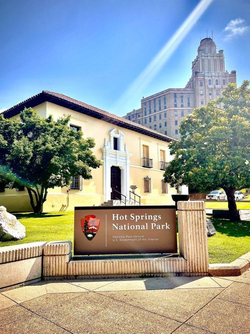 Sign for Hot Springs National Park in front of a historic building, with a tall structure visible in the background.