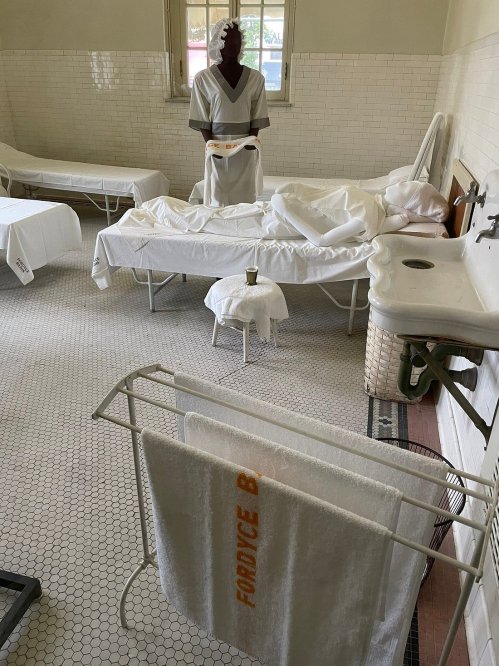 A historic medical room with white beds, towels, and a mannequin dressed in a hospital gown.