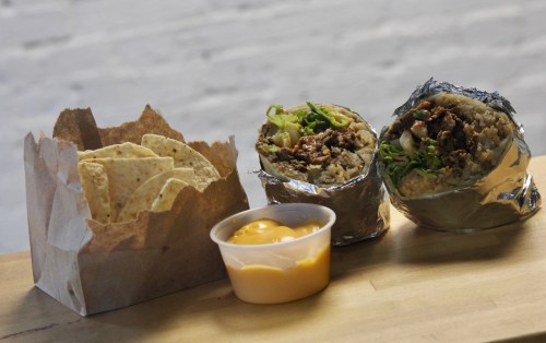 Two burritos wrapped in foil with green onions, alongside a container of cheese sauce and a box of tortilla chips.