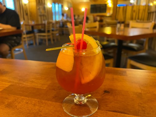 A colorful cocktail with fruit garnishes and two straws, sitting on a wooden table in a cozy restaurant setting.