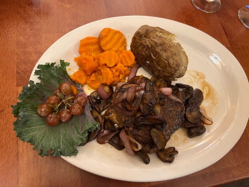 A plate featuring steak with mushrooms and onions, sweet potatoes, a baked potato, and a side of grapes and greens.
