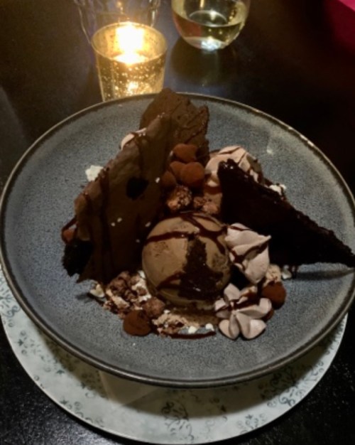 A dessert bowl featuring chocolate ice cream, chocolate shards, meringue, and toppings, with a candle in the background.