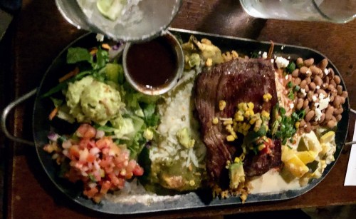 A sizzling platter with grilled meat, guacamole, salad, beans, pico de gallo, and a side of sauce.