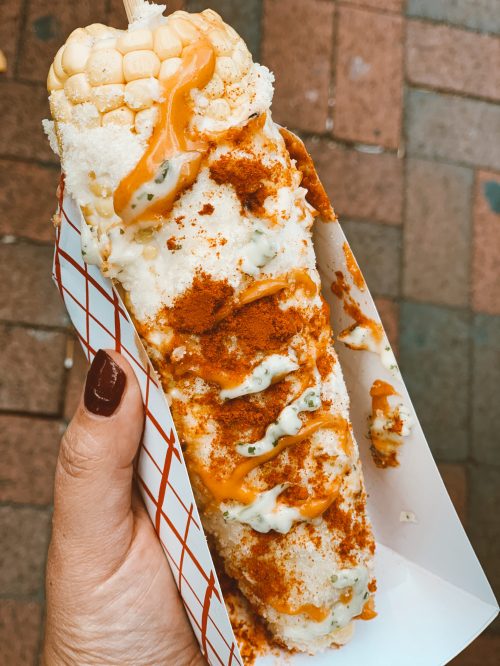 A hand holding a corn on the cob topped with mayonnaise, cheese, chili powder, and a drizzle of sauce.