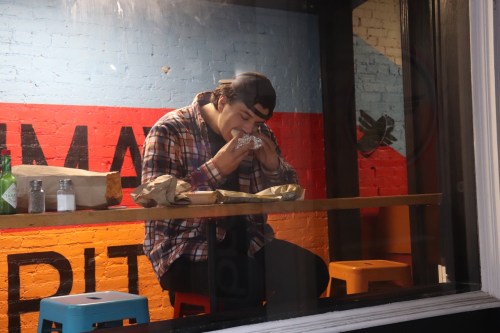 A person in a plaid shirt and cap sits at a table, eating a burrito inside a colorful restaurant.