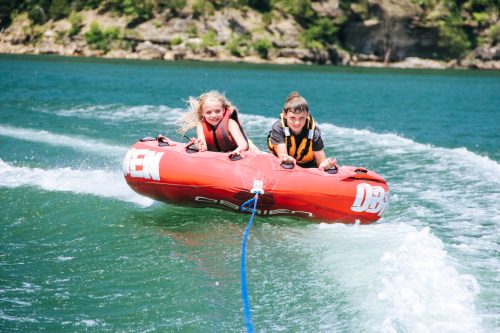 Two kids enjoy tubing on a lake, smiling as they glide over the water behind a boat.