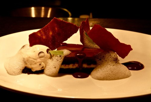A gourmet dessert featuring chocolate cake, foam, ice cream, and crispy red garnishes on a white plate.