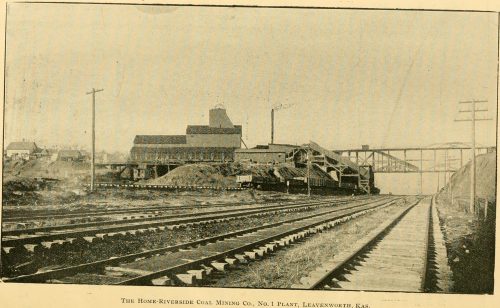 Black and white image of railway tracks leading to a building, with power lines in the background.