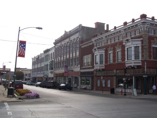 Historic downtown street with brick buildings, shops, and American flags hanging from streetlights.
