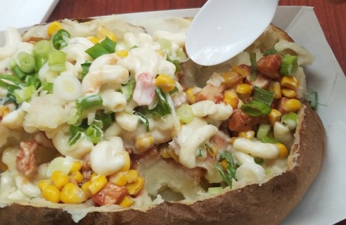 A baked potato topped with creamy macaroni, corn, green onions, and a drizzle of sauce.