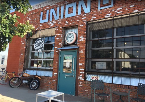 Exterior of a brick building with large windows, a clock, and a blue door. Bicycles and a motorcycle are nearby.