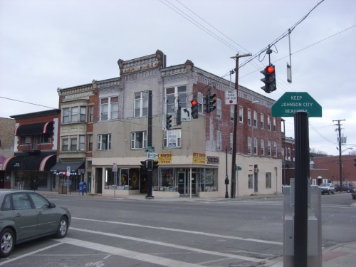 A street corner featuring a brick building with shops, traffic lights, and a directional sign for Johnson City.