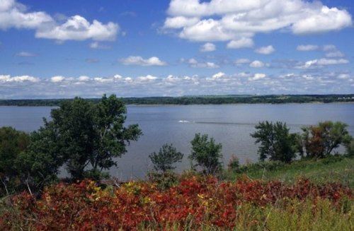 A serene river scene with lush greenery, colorful autumn foliage, and a blue sky dotted with fluffy clouds.