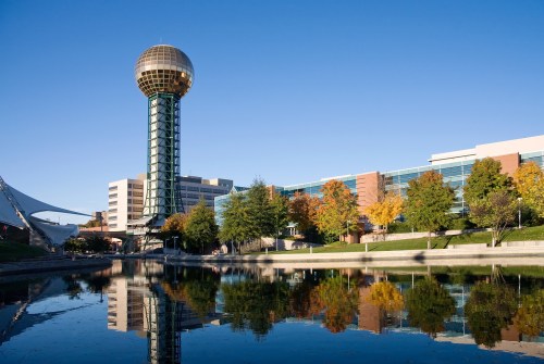A tall tower with a golden sphere on top reflects in a calm body of water, surrounded by colorful autumn trees.