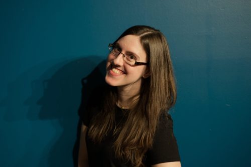 A smiling person with long hair and glasses stands against a blue wall, casting a shadow.