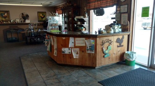A cozy café interior featuring a wooden counter, seating area, and decorative rooster artwork on the walls.