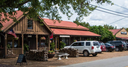 A rustic building with a red roof, flags, and parked cars, surrounded by trees and gravel.