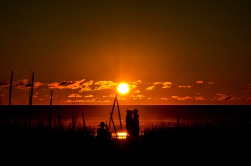 Silhouettes of people against a vibrant sunset over the ocean, with clouds and tall grass in the foreground.