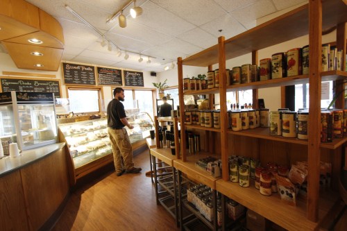 A cozy café interior with a display case, shelves of canned goods, and a customer browsing.
