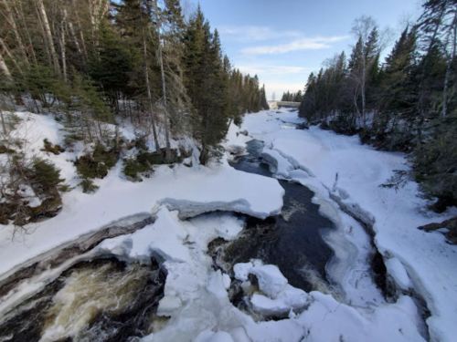 A snowy river landscape with ice formations and trees lining the banks under a clear blue sky.