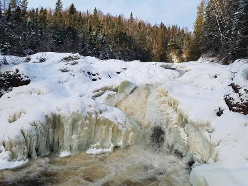 A frozen waterfall cascades over snowy rocks, surrounded by evergreen trees in a winter landscape.