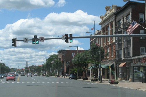 A street view with traffic lights showing green, lined with buildings and flags under a partly cloudy sky.