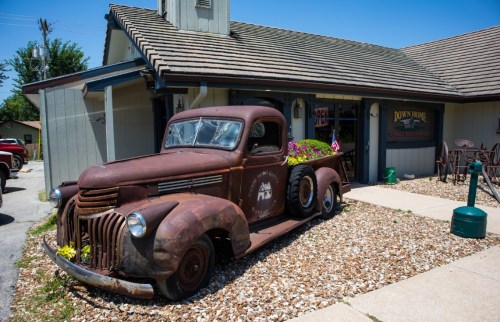 A vintage rusty truck with flowers parked outside a quaint building, surrounded by gravel and greenery.