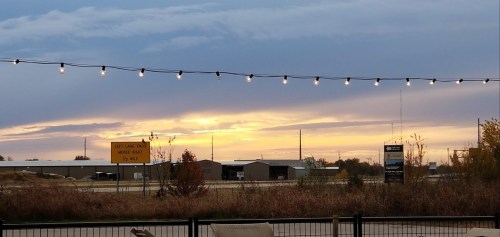 A sunset with colorful clouds over a rural landscape, framed by string lights and a roadside sign.