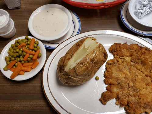 A plate with fried chicken, a baked potato, green peas, and carrots, alongside a bowl of creamy sauce.
